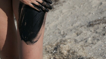 Healing black medical mud applies to leg and knee woman at the lake of salt water. Salty black dirt for people with health and skin problems. Relax at spa resort. Exfoliation scrub beauty treatment.