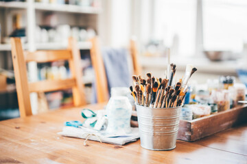 painting brushes of different form and size stand in small aluminium bucket on brown wooden table with various stuff of artist at workshop close view. - obrazy, fototapety, plakaty