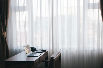 a notebook put on the wood table beside of the large window with curtain with nobody in the room of hotel or office in big city