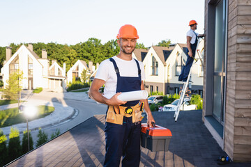 Selective focus of builder with toolbox and blueprint looking at camera while colleague working on...