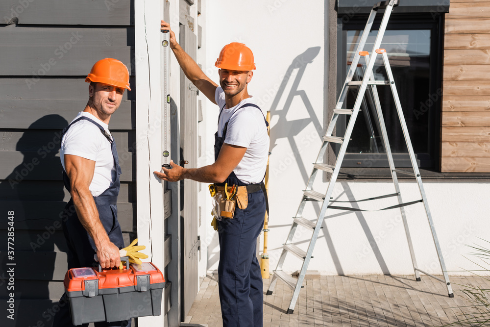 Wall mural Handymen with toolbox using spirit level and looking at camera near house