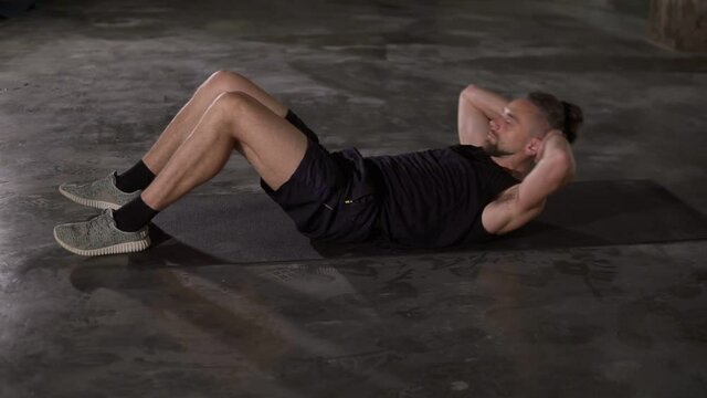 Muscular man in sportswear making abdominal sport exercise on fitness mat using alone in loft studio. Man doing abdominal fitness exercises in slow motion
