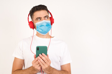 Fototapeta na wymiar Image of a happy young handsome man wearing medical mask posing isolated over bright background listening to music with earphones using mobile phone.