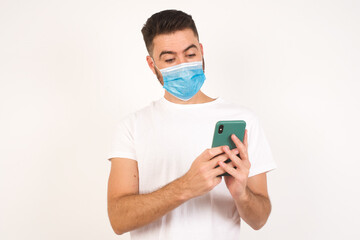 Fototapeta na wymiar Horizontal shot of handsome young male wearing medical mask, isolated over white background looks with bugged eyes, holds modern smart phone, receives unexpected message from friend, reads reminder.
