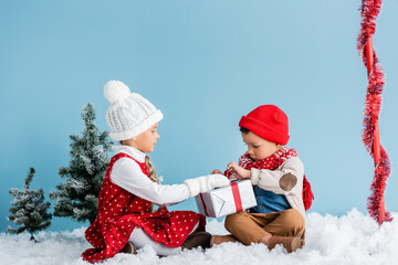 Obraz premium boy and girl in winter outfit sitting on snow and touching present isolated on blue