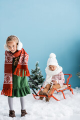 selective focus of girl in winter outfit giving a ride to brother on sleigh on blue
