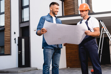 Workman in helmet holding blueprint near man and building outdoors