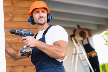 Selective focus of handyman in workwear and ear defenders holding electric screwdriver near facade...