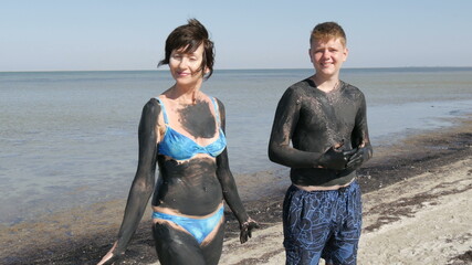 Oiled up mom and teen son smile Healing black medical mud at the lake of salt water. Salty black dirt for people with health and skin problems. Relax at spa resort. Exfoliation scrub beauty treatment.