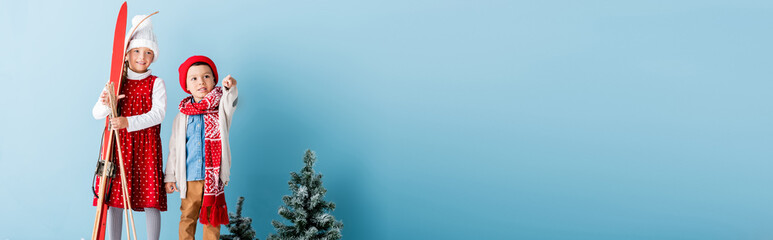 panoramic crop of boy in hat and scarf pointing with finger near sister holding ski poles and skis while standing on blue