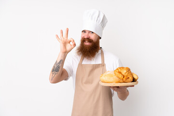 Redhead man in chef uniform. Male baker holding a table with several breads showing ok sign with...