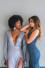 Two multi-ethnic friends in ball gowns ready to go out at night to the exclusive party. Gray and blue evening dresses, blonde caucasian girl, black girl with afro hair, gray wall background