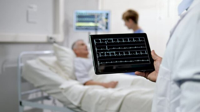 Close up on doctor hand holding tablet showing livestream ecg electrocardiogram with patient lying on hospital bed in the background