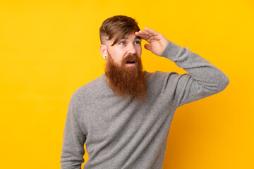 Redhead man with long beard over isolated yellow background looking far away with hand to look something