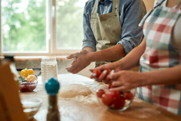 Cropped shot of couple making pizza together. Young man in apron making the dough while woman preparing tomatoes. Hobby, lifestyle