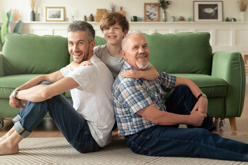 Happy grandson embracing young handsome father and grandfather looking at camera laughing together at cozy home. 
