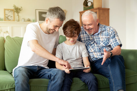 Handsome young man sitting on couch with smiling aged father and schoolboy son, using funny tablet apps together. 