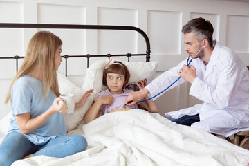 Obraz premium Doctor with stethoscope listening to lung sound of little girl laying in bed looking at her anxious mother.