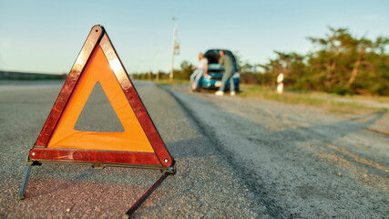 Red warning triangle sign on the road with a couple checking, standing near their broken car in the background