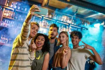 Young men and women smiling while taking selfie using smartphone. Multiracial group of friends hanging out at party in the bar