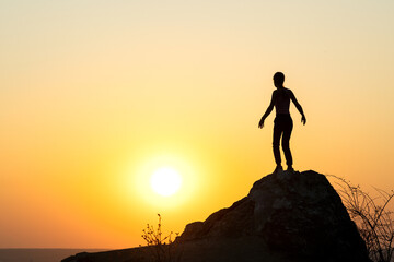 Fototapeta premium Silhouette of a woman hiker standing alone on big stone at sunset in mountains. Female tourist on high rock in evening nature. Tourism, traveling and healthy lifestyle concept.