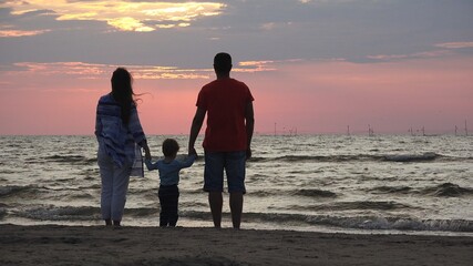 Parents holding child standing on seashore, family admire sunrise
