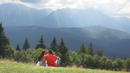 Portrait of couple of lovers sitting with back admiring gorgeous mountains peak landscape, sun rays shining through white clouds, peace in the heart of nature