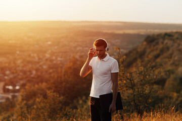 Young man standing on a hill and holding the laptop. Young hipster man with headphones listening the music with orange sunset on background. 