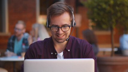 Happy guy with wireless headset having online conference at cafe using laptop