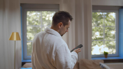 Confident young man walking out bathroom in robe after shower checking messages on smartphone