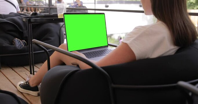 Young Woman at Coffee Shop Working on a Laptop MacBook Pro Computer with Green Mock-up Screen. She's Sitting On a Bean Bag Chair in Coworking open space outdoor office. Over the Shoulder Camera Shot.