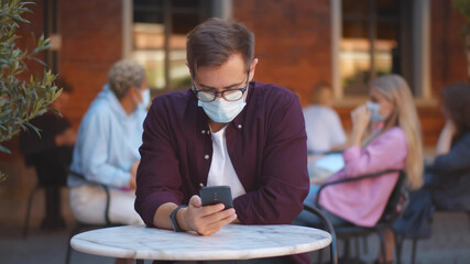 Young guy wearing facial mask surfing internet on mobile phone in outdoors cafe