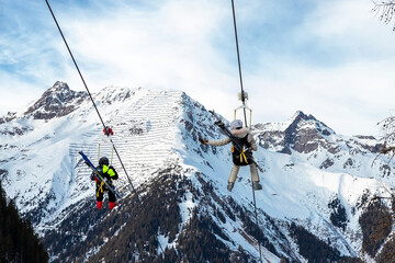 Scenic view couple of tourist with skiing equipment enjoy having fun riding extreme suspended zipline wire flying over gorge canyon against snowcapped mountains on background. Adrenaline recreation