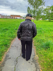 old man with a cane and a black jacket with a black hat walks along the sidewalk rear view