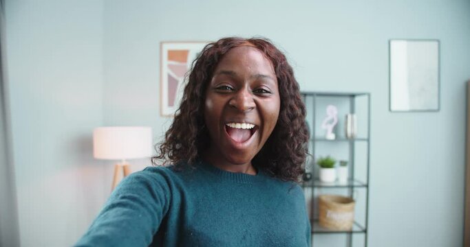 POV of happy African American girl talking on video call in good mood at home while sitting down on couch. Joyful beautiful female videochatting in room with smile on face. Conversation concept