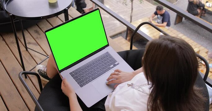 Girl using keyed blank green screen MacBook Pro in outdoor cafe food market. Travel freelance business lifestyle. Internet communication chat. Typing message, searching. Chromakey mockup. Top view