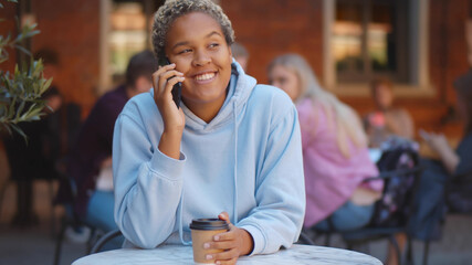 Young african woman talking on smartphone drinking takeaway coffee at table outdoors