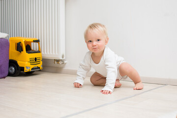 One year old blond baby boy sitting on floor at home and looking at camera. Little kid is crawling on all four