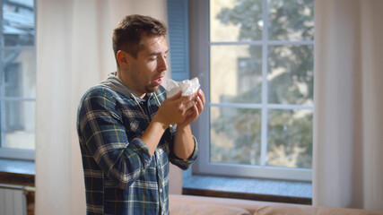 Sick man blowing his runny nose in paper tissue walking at home