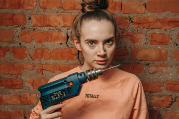 girl with a drill, against the background of the wall, looks at the camera and makes repairs