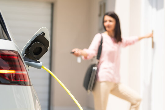 Close up of a electric car charger with female silhouette in the background, entering the home door and locking car