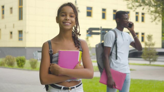 Portrait of charming African American schoolgirl posing outdoors with her busy brother talking on the phone at the background. Smiling pretty girl standing on college yard with workbook and backpack.