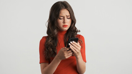 Upset offended girl in red top disappointedly using smartphone over white background