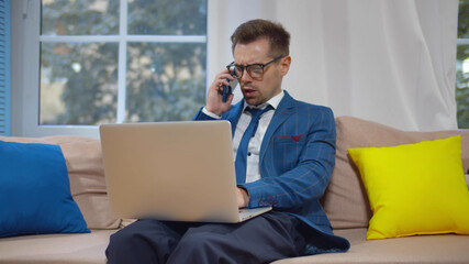 Businessman talking on mobile phone working on laptop sitting on couch.