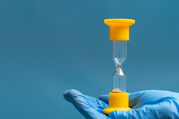 A hand in a blue protective glove holds an hourglass on a blue background, close-up, copy space