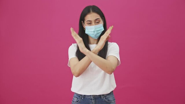 A pretty young woman with protective mask is showing stop gesture while crossing her arms standing isolated over a pink background