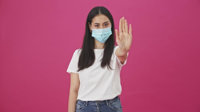 A serious young woman with protective mask is showing stop gesture standing isolated over a pink background