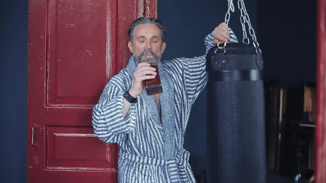 Drunk bearded man ignores sports. He stands in a robe with a bottle of whiskey, leaning on a boxing pear. Drinks alcohol and strokes a boxing pear. High quality FullHD footage.