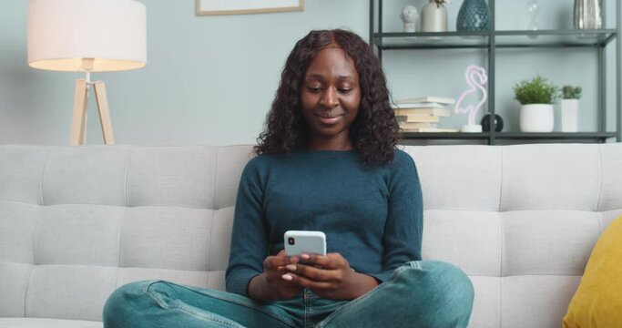 Portrait of happy beautiful girl texting on smartphone and laughing while sitting on couch at home. Joyful African American young pretty female typing on mobile phone in room. Rest concept