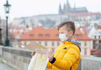 Young boy wearing medical mask holds map against the background of Prague Castle. Coronavirus epidemic concept. Empty space for text